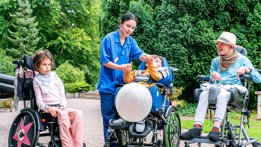 A caregiver interacts with three children in wheelchairs outdoors, engaging them with a large balloon. Trees and greenery in the background.