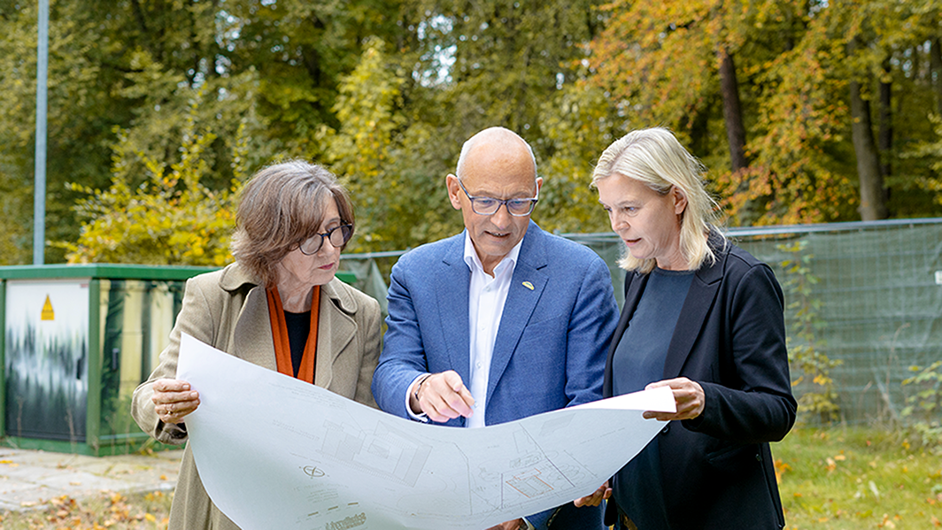 Ein Mann und zwei Frauen stehen draußen und beratschlagen vor einem großen Bauplan.