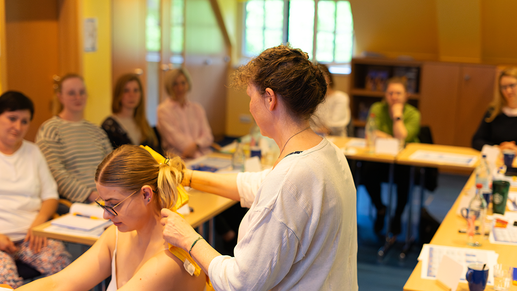 Mehrere Personen sitzen an Tischen in einem Seminarraum. Eine Frau steht und legt einer sitzenden Frau ein gelbes Handtuch um den Nacken.
