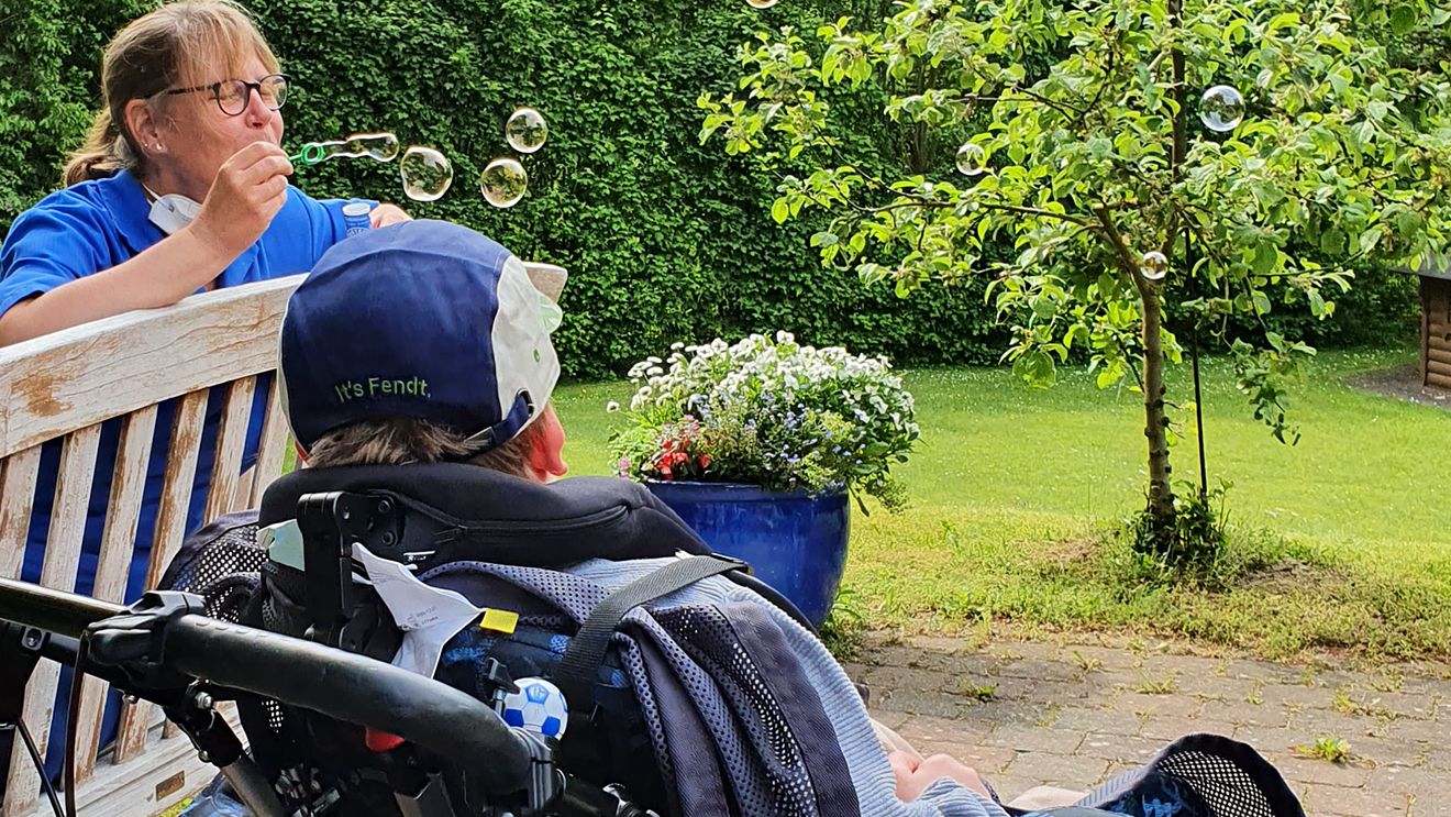 A caregiver blows bubbles near a person in a wheelchair in a garden, with a tree and flowers in the background.