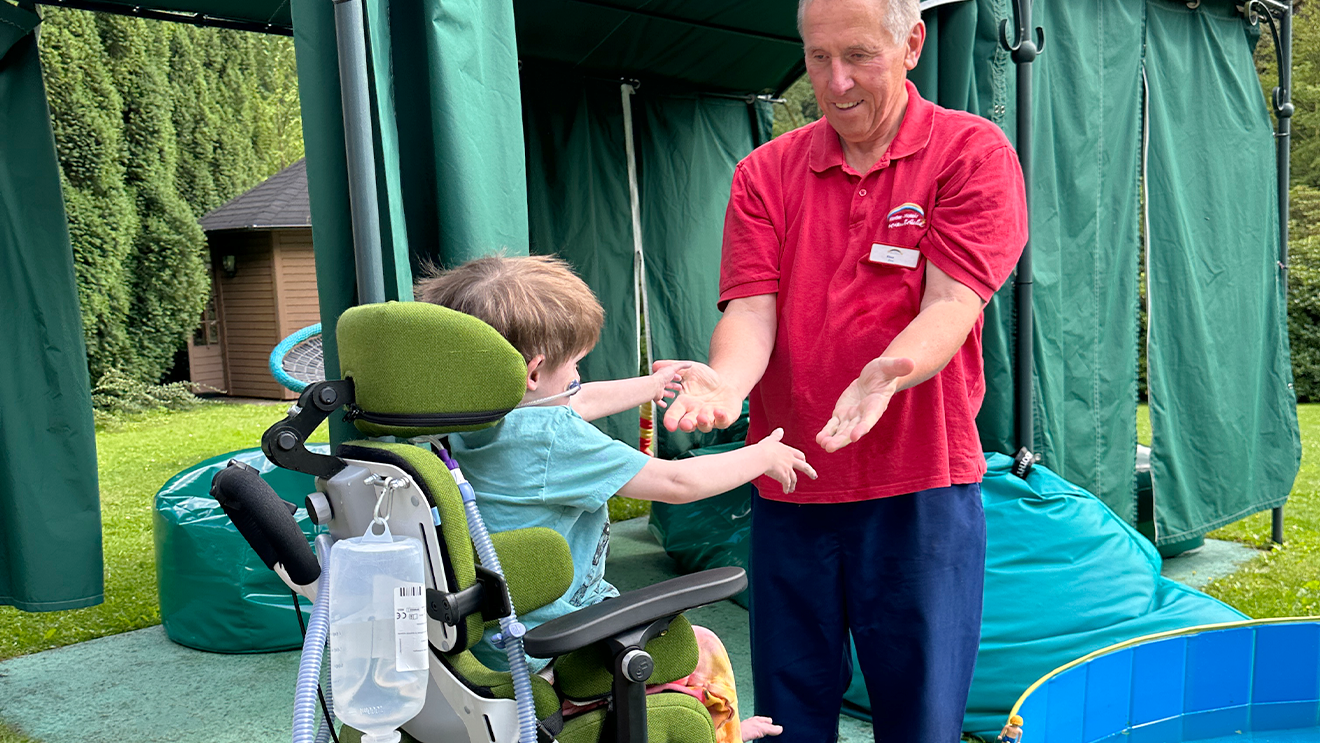 A man in a red shirt helps a child in a green wheelchair reach out, set outdoors with green structures and bean bags in the background.