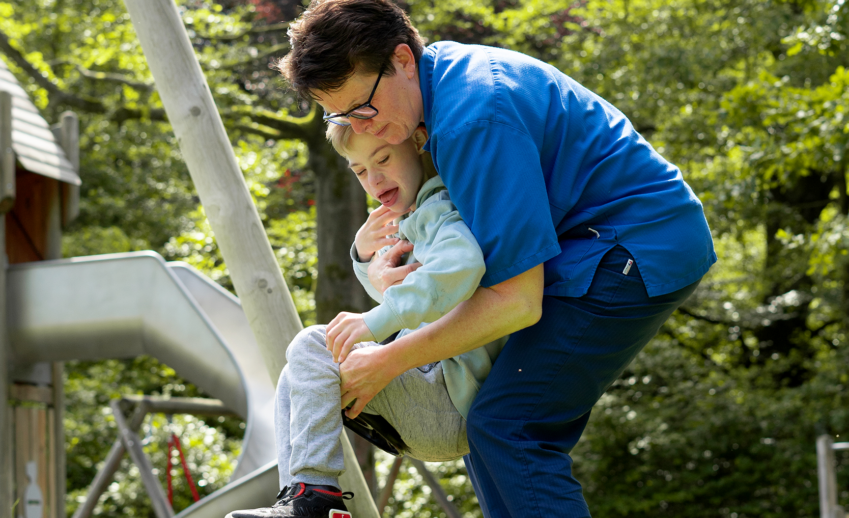 Eine Betreuerin in blauer Kleidung hilft einem Kind auf einer Schaukel in einem sonnigen Spielplatz mit einer Rutsche und Bäumen im Hintergrund.