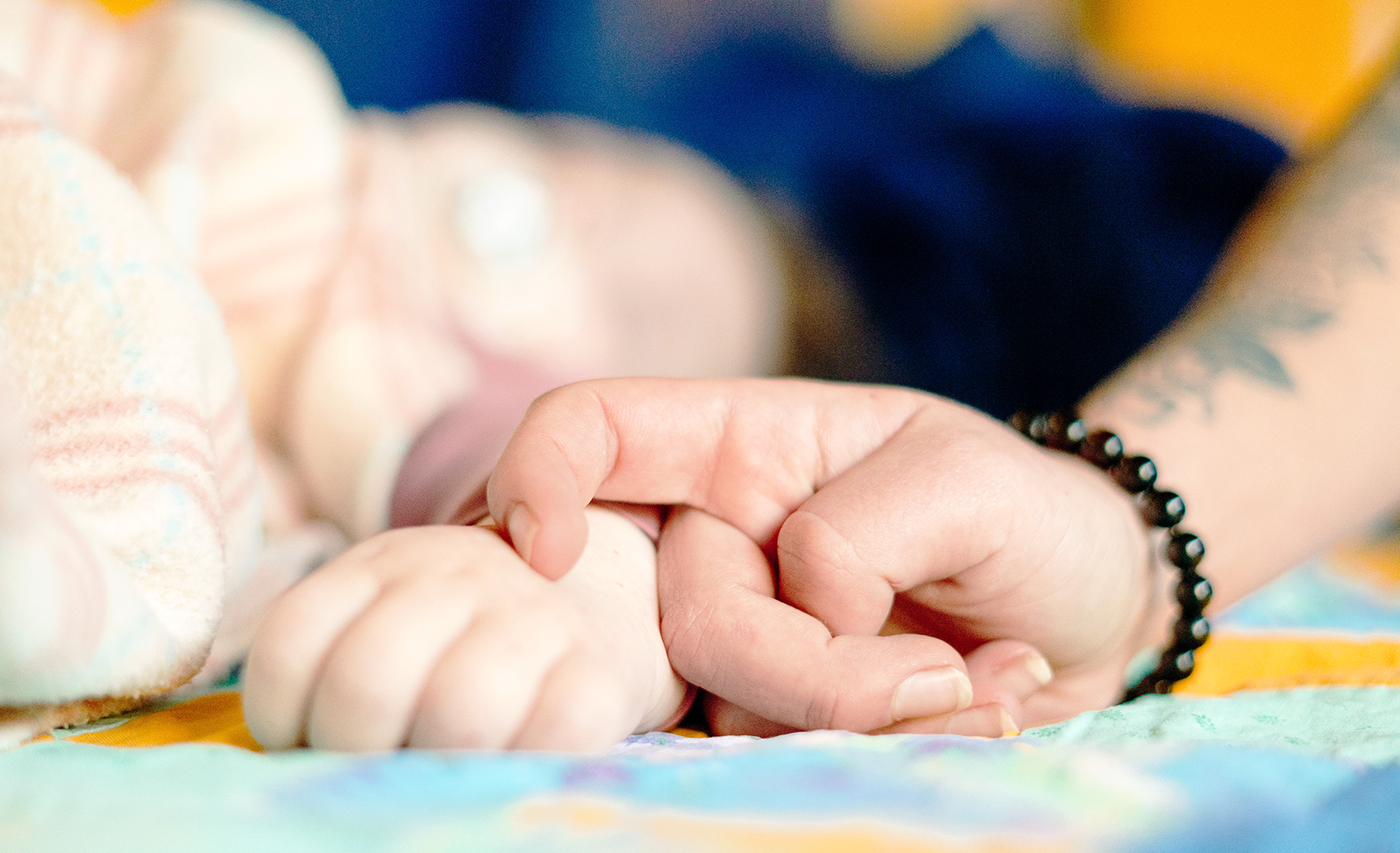 An adult’s hand touches a child’s hand. Both are lying on a colorful blanket. The adult’s arm is tattooed and wears a black beaded bracelet.