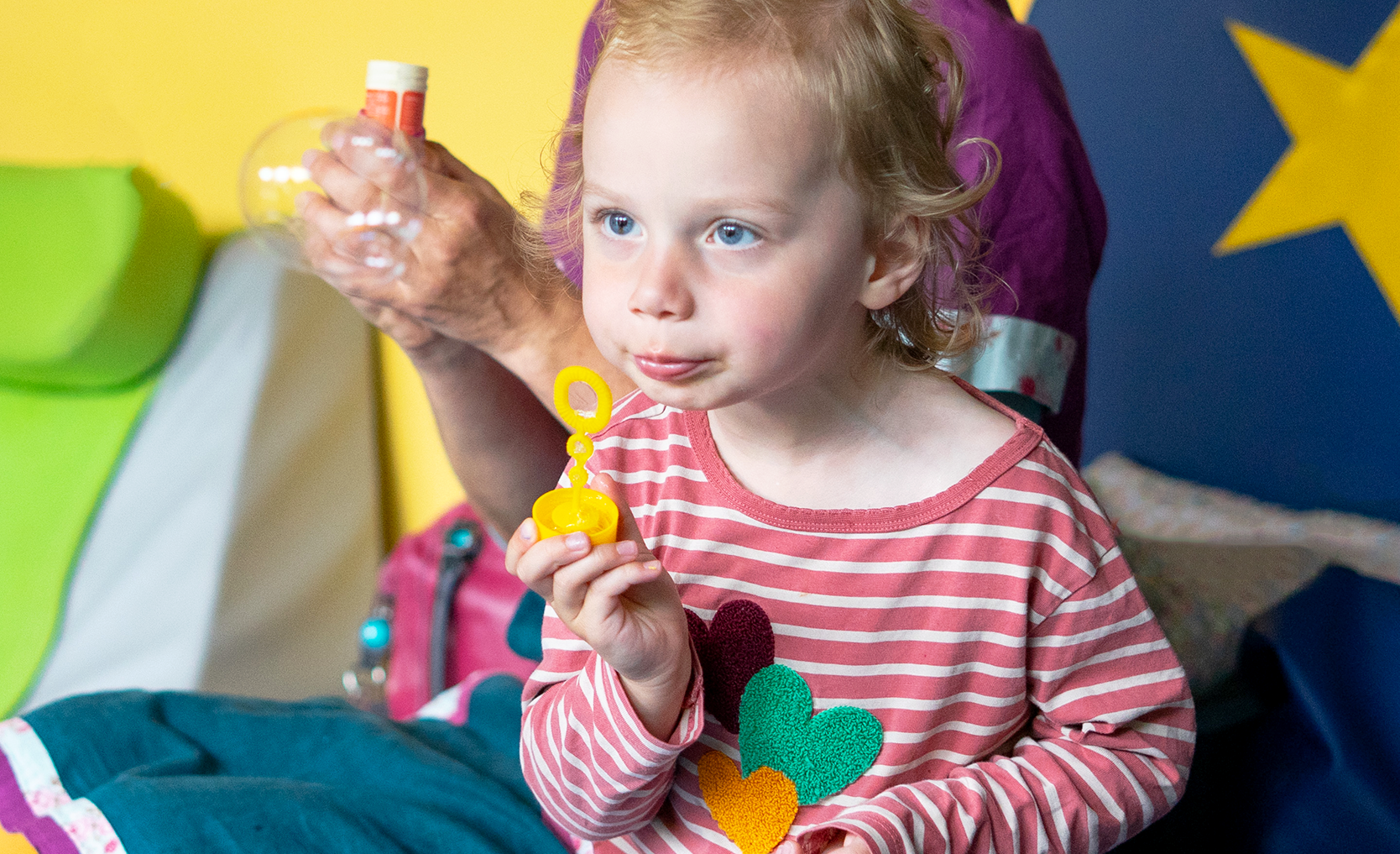 A young child with curly hair blows bubbles, wearing a pink striped shirt with colorful heart shapes. A person in the background holds a bubble bottle.