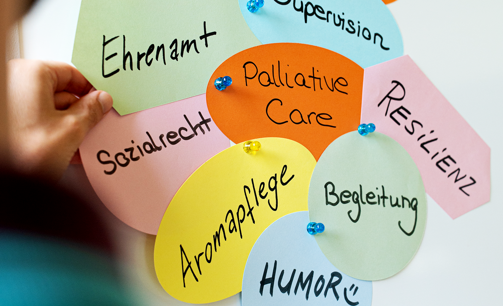 Bunte Papierformen mit handgeschriebenen deutschen Wörtern wie „Ehrenamt“, „Palliative Care“ und „Resilienz“, die an eine Tafel geheftet sind.