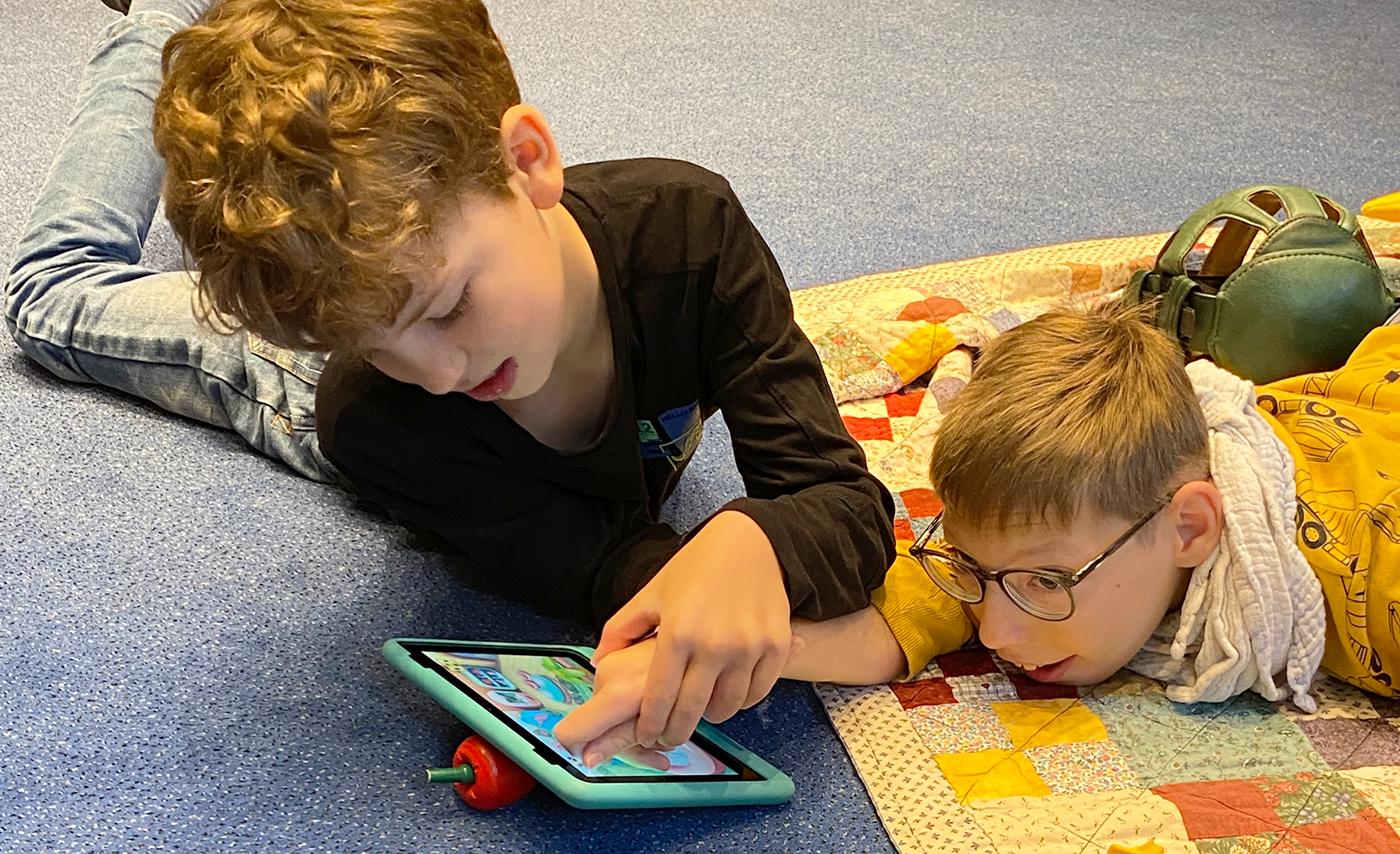 Zwei Jungen liegen auf einer gesteppten Decke und sind auf ein Tablet konzentriert. Einer zeigt auf den Bildschirm, beide sind mit der Aktivität beschäftigt.