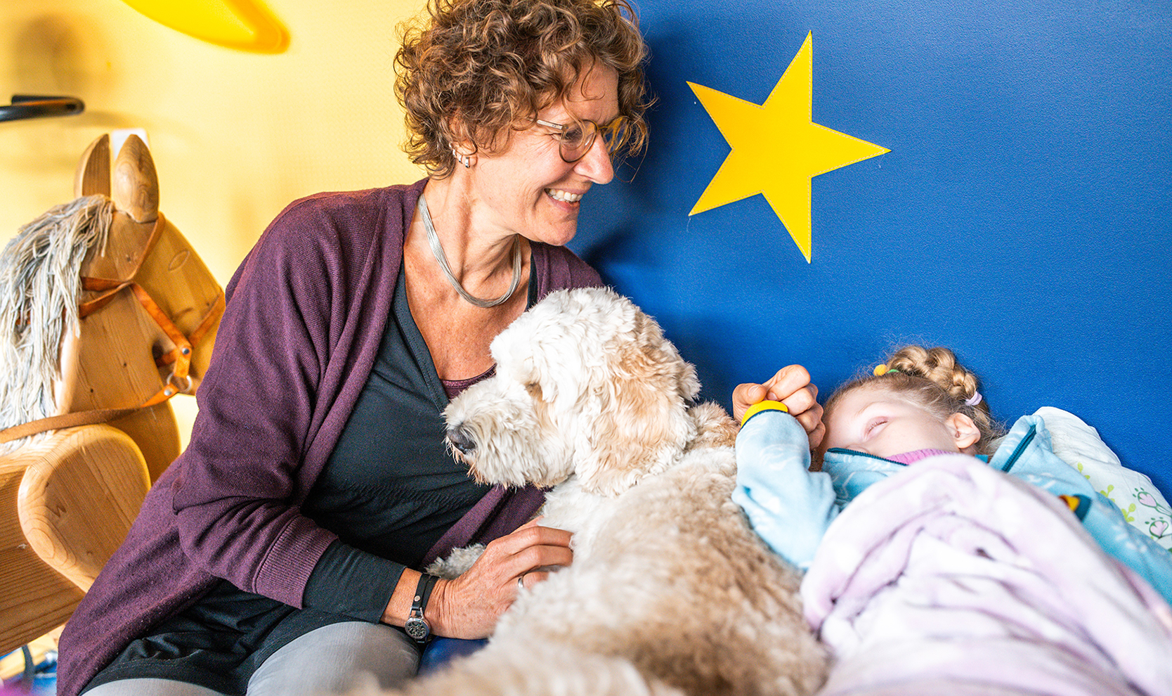 Eine Frau mit lockigem Haar hält sanft die Hand eines schlafenden Mädchens. Ein Hund liegt zwischen ihnen.