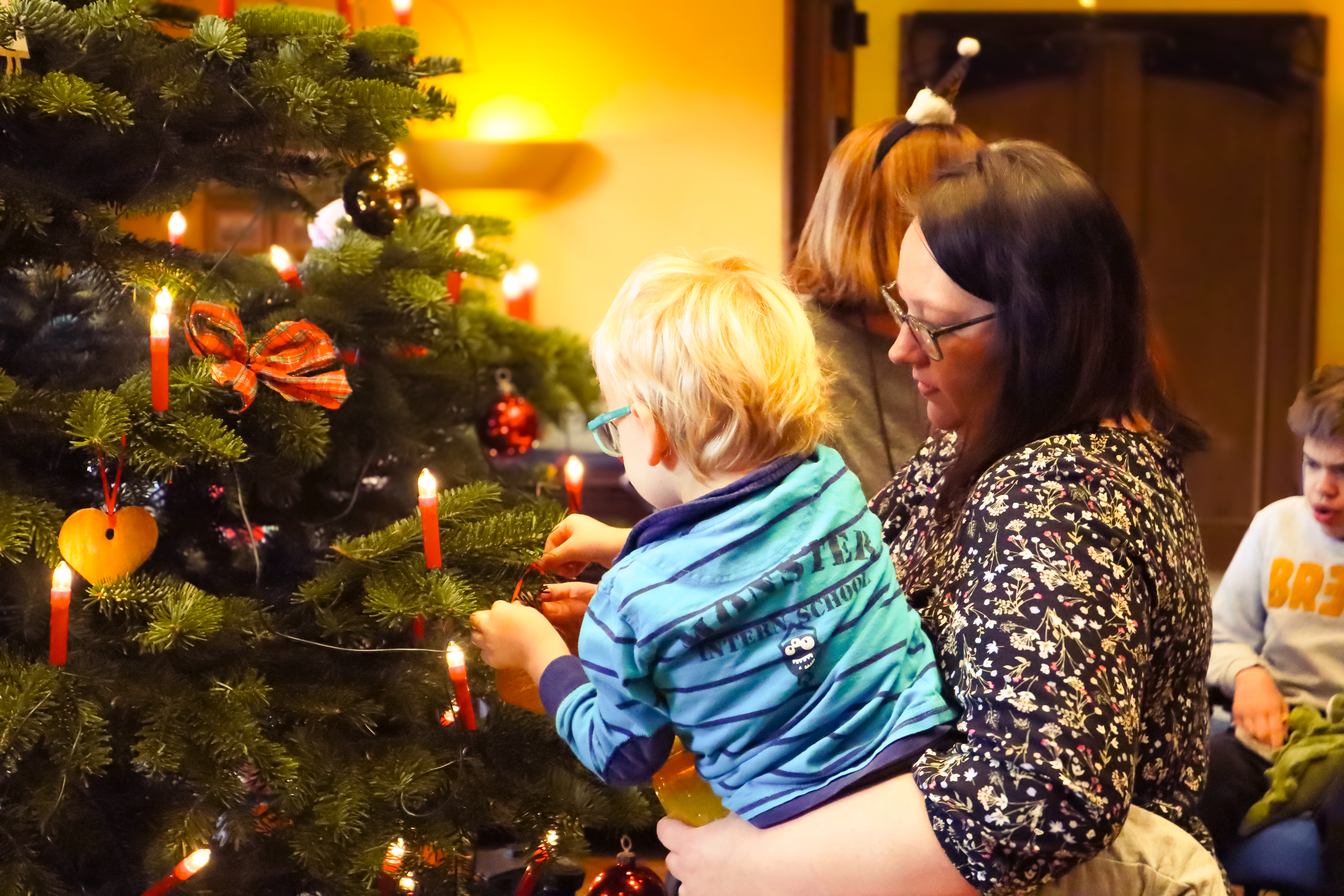 Eine Frau hält ein Kind im Arm, während sie einen Weihnachtsbaum mit roten Kerzen und Christbaumschmuck schmücken. Ein weiteres Kind sitzt in der Nähe und trägt einen festlichen Pullover.