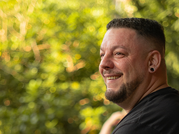 Ein lächelnder Mann mit kurzen Haaren und Ohrringen steht im Freien vor einem unscharfen grünen Hintergrund.