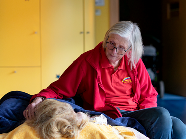 Eine ältere Frau in einer roten Jacke sitzt neben einer Person, die sich ausruht, und berührt sanft deren Kopf in einem gemütlichen Raum mit gelben Schränken.