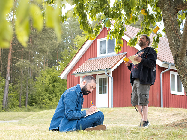 Zwei Männer machen sich Notizen unter einem Baum in der Nähe eines roten Hauses mit weißer Verzierung, umgeben von Grün.
