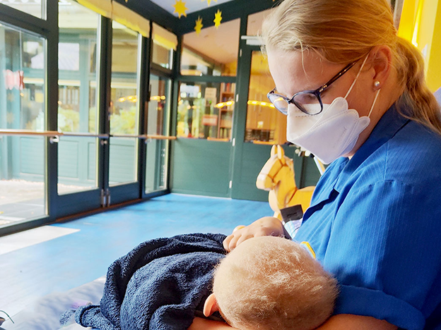 A caregiver in a mask holds a child wrapped in a towel in a brightly lit room with large windows and playful decor.