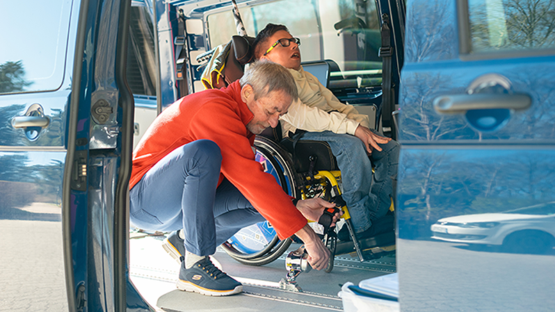 A person assists a young wheelchair user in a vehicle, adjusting the wheelchair's securement.