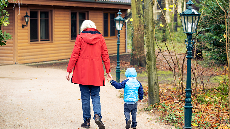 Eine ältere Frau mit roter Jacke läuft zu einer Blockhütte und hat einen kleinen jungen in Winterjacke an ihrer Hand.