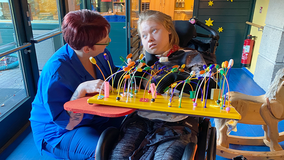 A caregiver interacts with a child in a wheelchair. The child is engaged with a colorful sensory board.